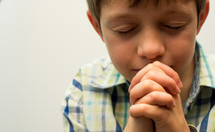 A boy folds his hands in prayer  with his eyes closed.