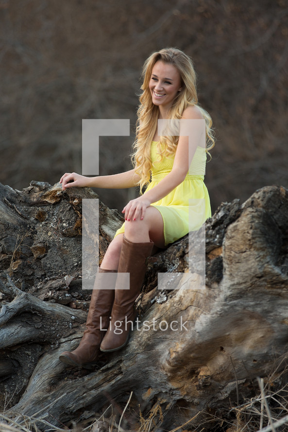 A woman in boots and a yellow dress sitting on a rock outdoors. 
