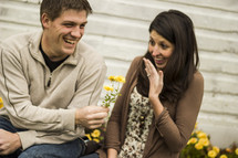 Boy and girl sitting by flowers and a building; he's giving her flowers; she's showing off her engagement ring