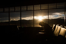 Silhouette of a man sitting by an airport window n prayer at sunrise.