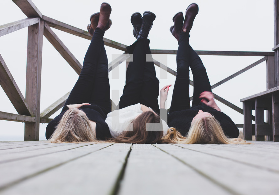 Three women laying on their backs on a deck, with their feet up on the railing.