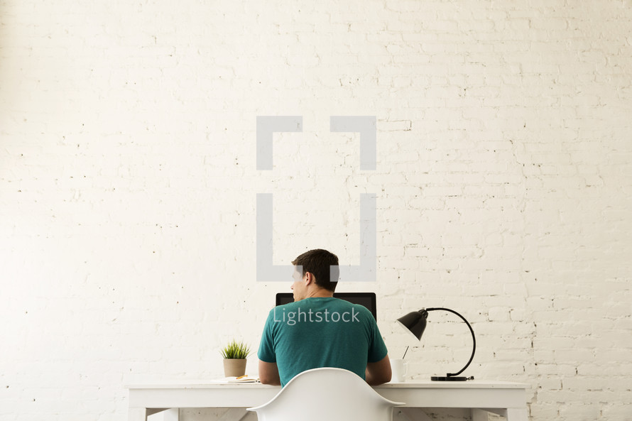 Man sitting at a computer desk against a white brick wall.