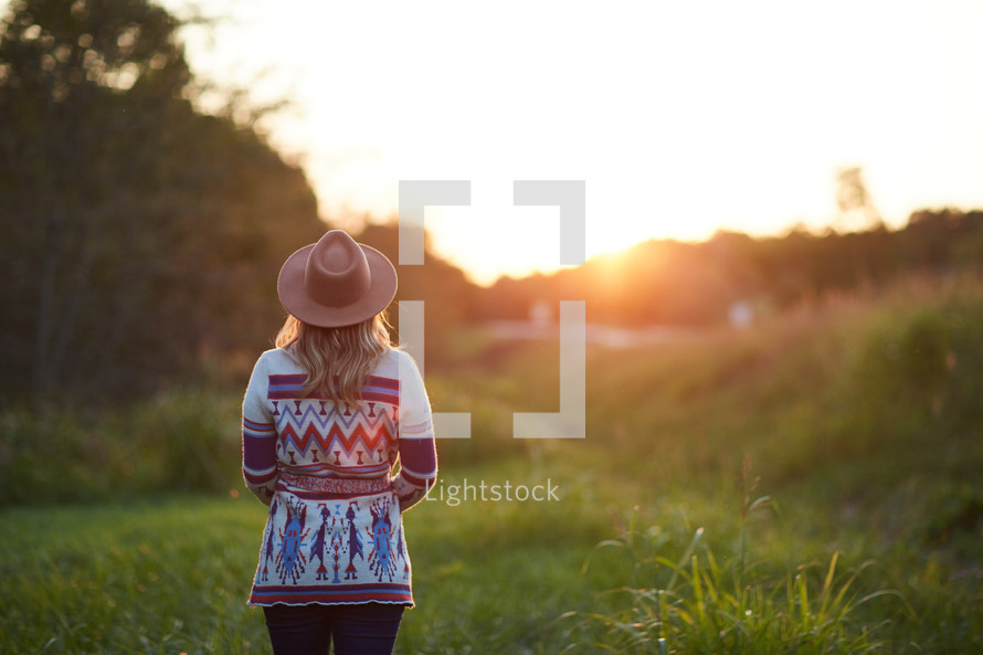 a woman in a sweater and hat standing in the middle of a field at sunset 