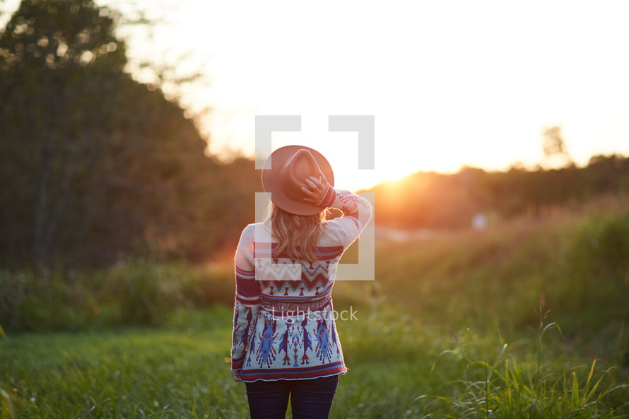 a woman in a sweater and a hat standing in the middle of a field at sunset 