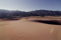 Person walking through sand in the desert, leaving a trail of footprints, with mountains in the background.