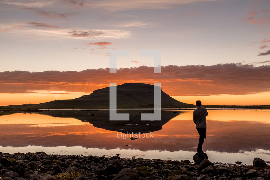 man standing on a rocky shore and reflection of a rock peak on calm water at sunset 