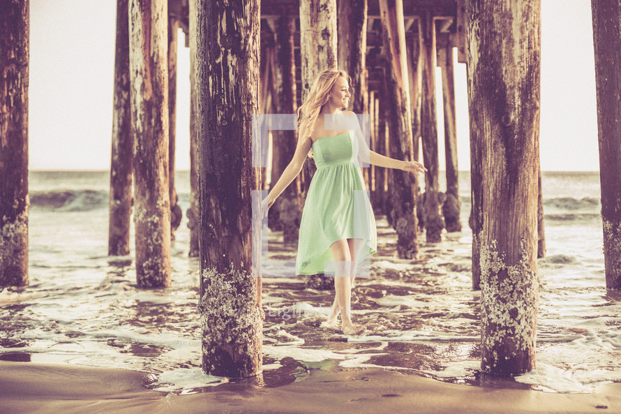 A woman walking in the water under a pier at a beach.