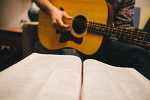 A man playing a guitar while looking at a Bible. 