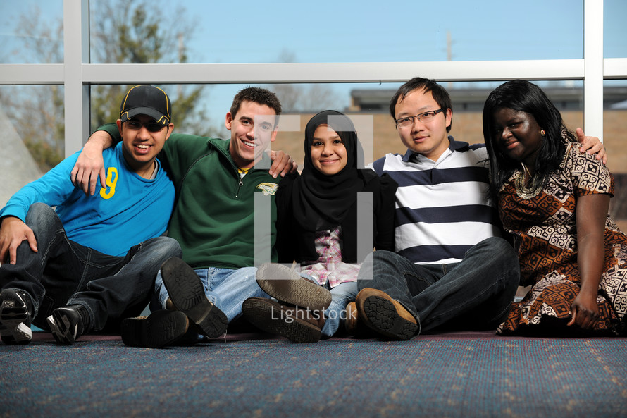 Five young people of different ethic backgrounds, arms around each other, sitting on a floor outdoors
