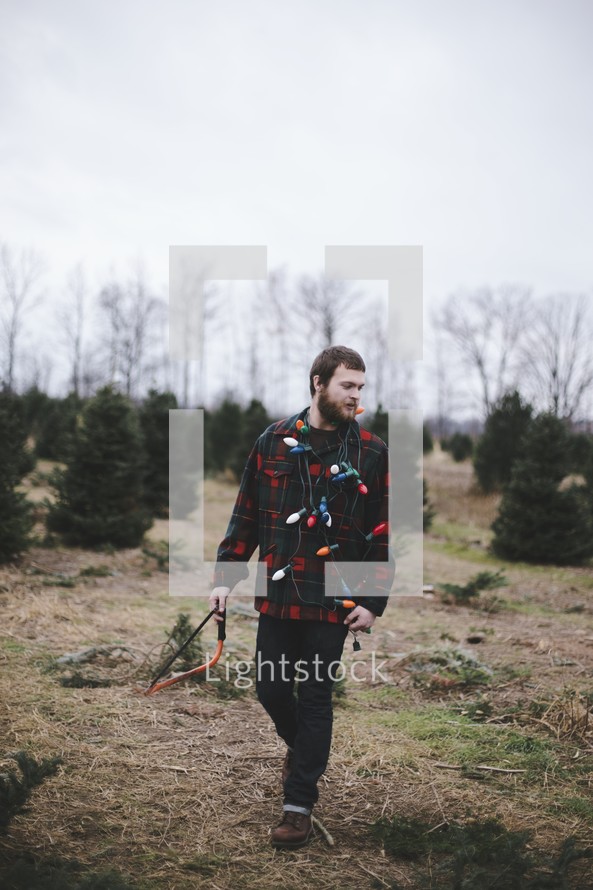 a man walking with a saw and Christmas lights in a Christmas tree lot 