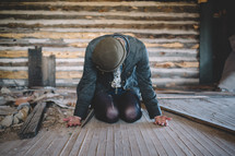 A woman sits bent over on the floor of an abandoned log house.