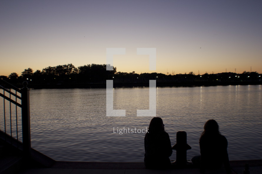 silhouettes of girls sitting on a dock looking out at a lake at dusk 