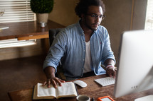 An African American man sitting at a desk typing on a computer and reading a Bible 