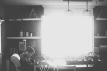 Brilliant sunlight fills a kitchen where a boy sits at the table with his head bowed.