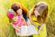 mother and daughters reading a Bible together outdoors