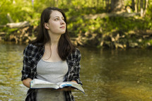 A woman standing near a stream with an open Bible, looking skyward.
