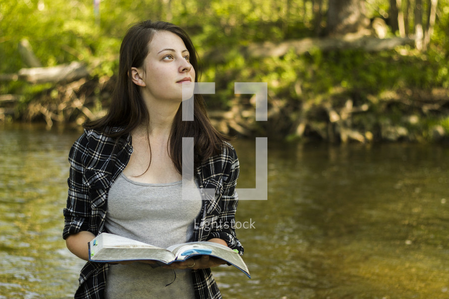 A woman standing near a stream with an open Bible, looking skyward.