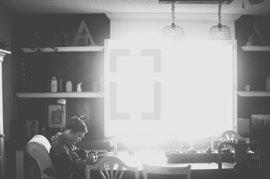 Brilliant sunlight fills a kitchen where a boy sits at the table with his head bowed.