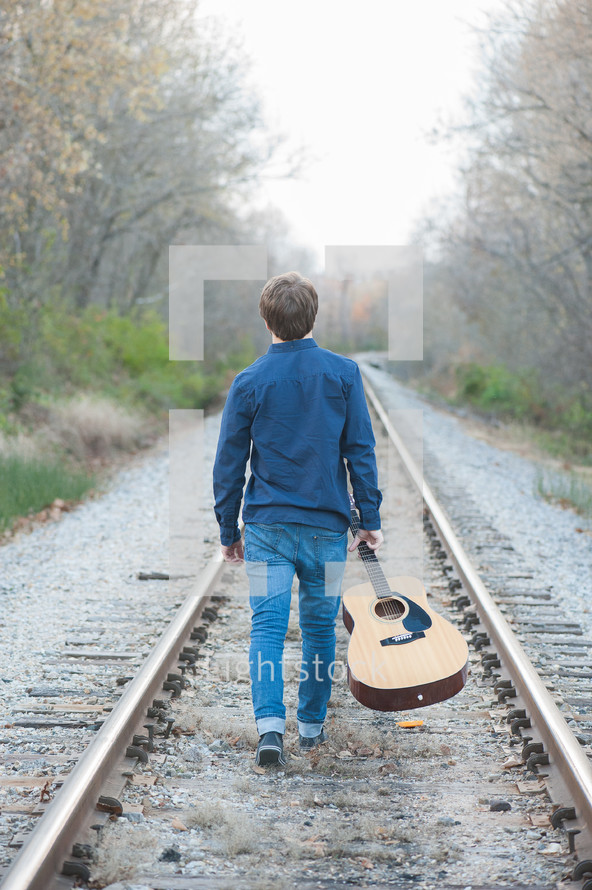 Man walking on a railroad track holding an acoustical guitar.