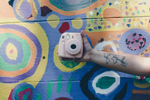 arm with a world map tattoo holding a camera in front of a painted wall 
