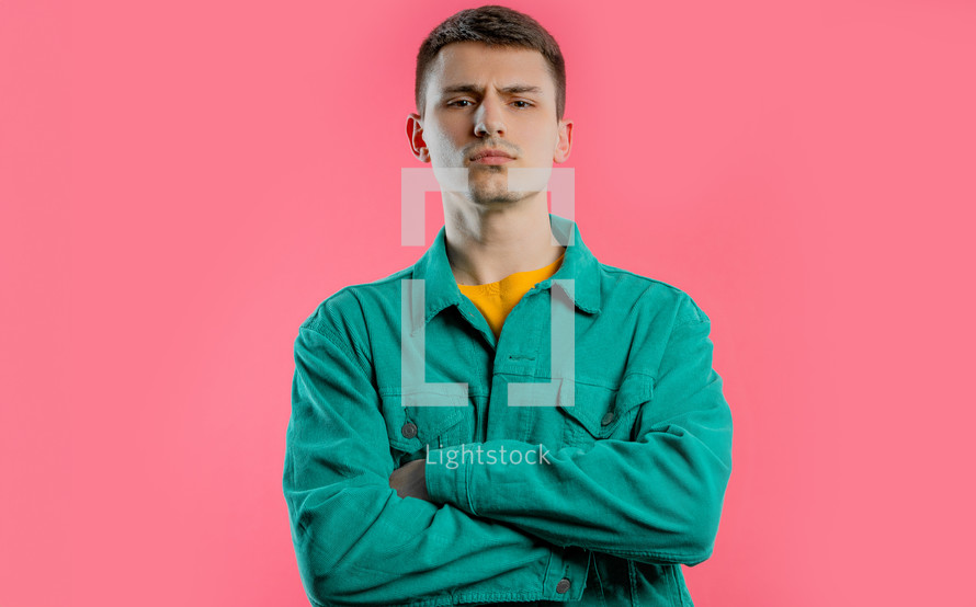 Offended upset young guy with arms crossed feeling mad at someone. Teen maximalism. Facial expressions, emotions and feelings. Body language. Self-confident man on pink background.