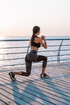 Young athletic woman performs squats on wooden embankment by the sea in early morning. Healthy lifestyle, coaching, training concept
