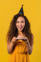 Happy birthday woman making wish - candle on cake. Girl smiling, celebrating anniversary. Young stylish lady on yellow background. High quality photo