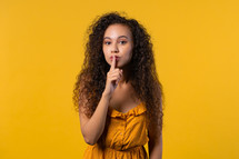 Smiling ukrainian woman holding finger on lips, yellow studio background. Pretty lady with gesture of shhh, secret, silence, conspiracy, gossip concept. High quality
