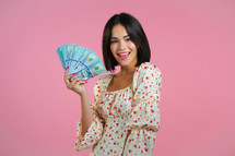 Amazed happy excited woman showing money - U.S. currency dollars banknotes on pink wall. Symbol of success, gain, victory