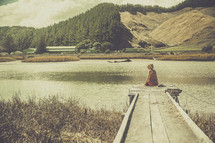 A woman sitting at the end of a wooden pier and looking at a lake.