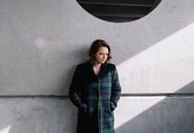 a woman in a plaid coat leaning against a concrete wall 