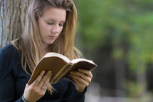 Girl sitting outside, leaning against a tree, reading a bible.