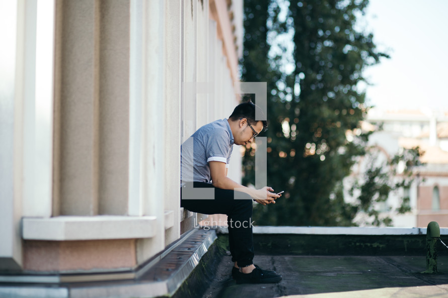 a man sitting on a rooftop texting on his cellphone 