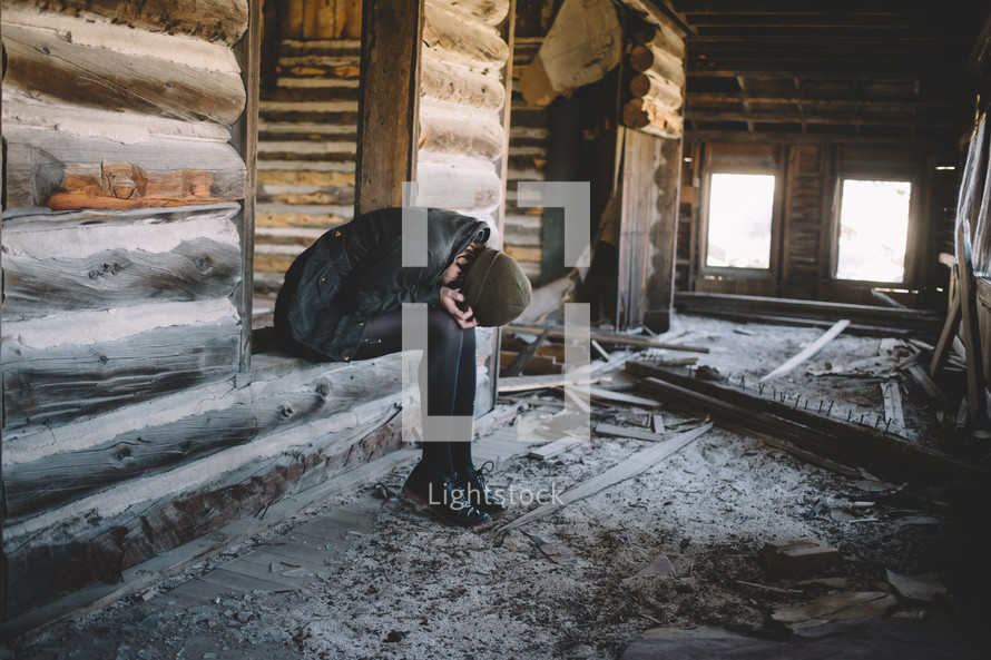 A woman bent over in grief in an abandoned log house.