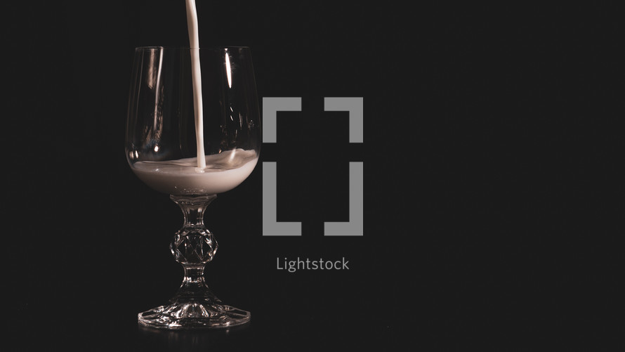 A transparent glass for wine is filled with milk on a black background