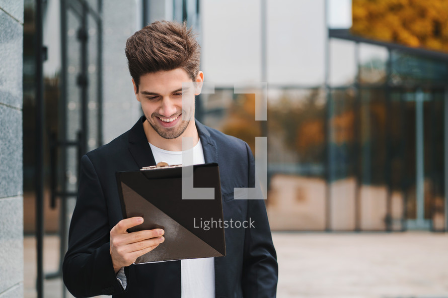 Portrait of young handsome man in jacket with documents, utility bills, report. Businessman near office building. He is satisfied with work of corporate staff. High quality photo