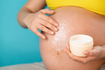 Close up of pregnant woman applying moisturizing cream against stretch marks on belly with baby on blue background. Maternity, motherhood, pregnancy, love concept
