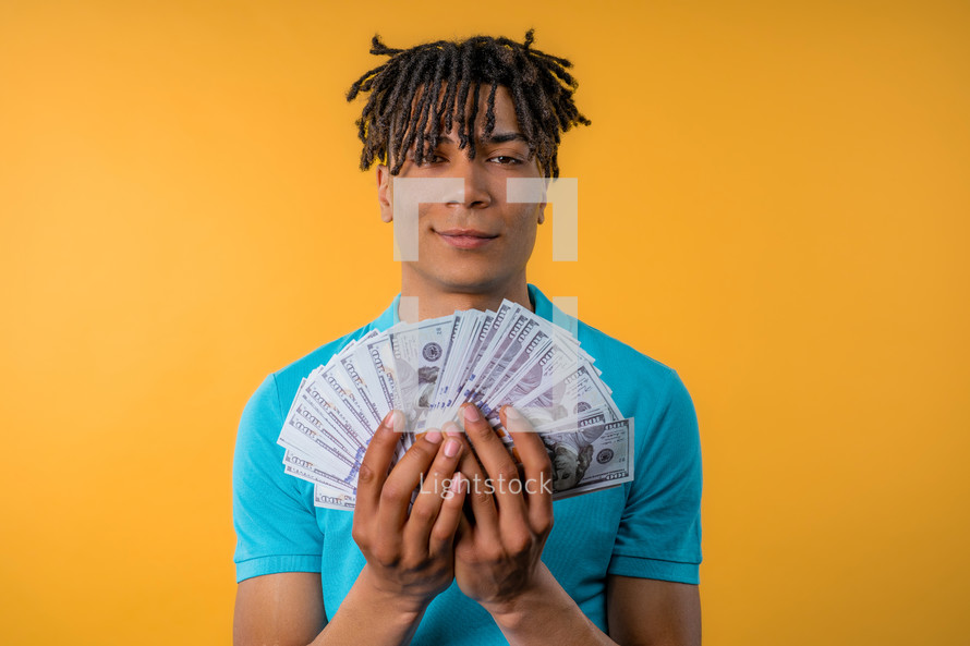 Satisfied man with USD currency. Young millenial african american guy or student holding money - dollars banknotes on yellow wall. Symbol of success, gain, salary, benefit, investments. 