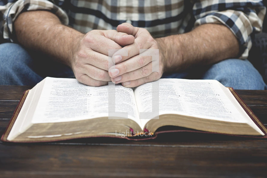 a man in a plaid shirt with praying hands over the pages of a Bible 