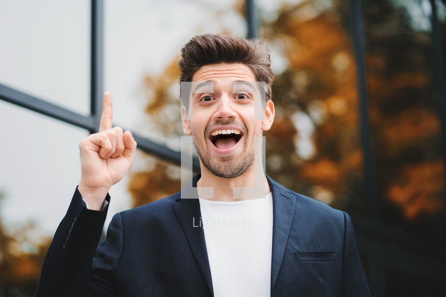 Smiling happy student man showing eureka gesture. Portrait of young thinking pondering businessman having idea moment pointing finger up on office building background