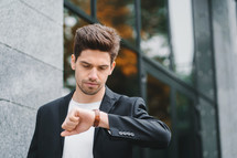 Handsome businessman or student looks at watch. Young man in hurry late for work. Male model on office building background.