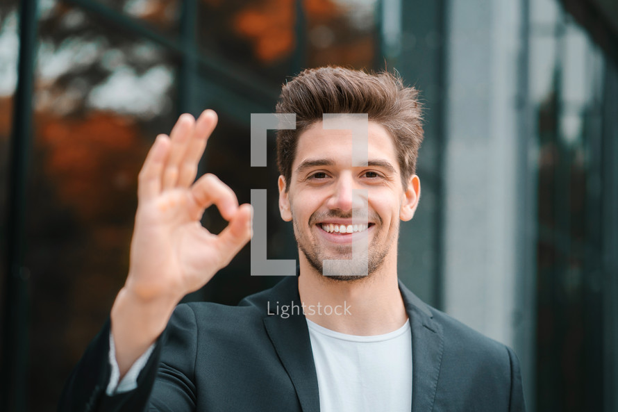 Successful happy man looking to camera and showing OK sign on office building background. Professional male manager wearing corporate suit jacket. Student guy outdoors.