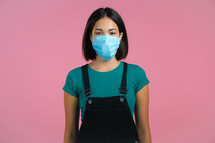 Young pretty girl in face medical mask during coronavirus pandemic. Portrait on pink background. Protection with respirator against COVID-19 outbreak.