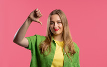 Portrait of unhappy european woman condemns with sign of dislike. Young millennial lady expressing discontent with showing thumbs-down gesture on pink studio background.