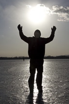 Silhouette of man praising God, hands raised and standing on ice at sunset.