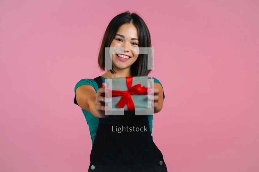 Excited woman holding gift box and gives it by hands to camera on pink wall background. Girl smiling, she is happy with present. Studio portrait.