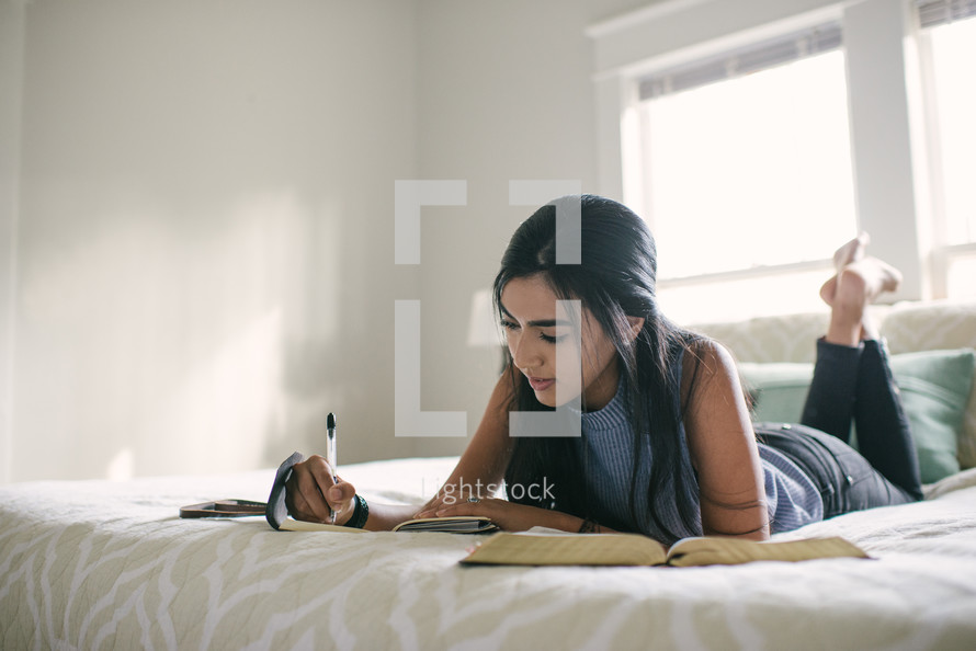 A teen girl taking notes and reading the Bible while laying on her bed.