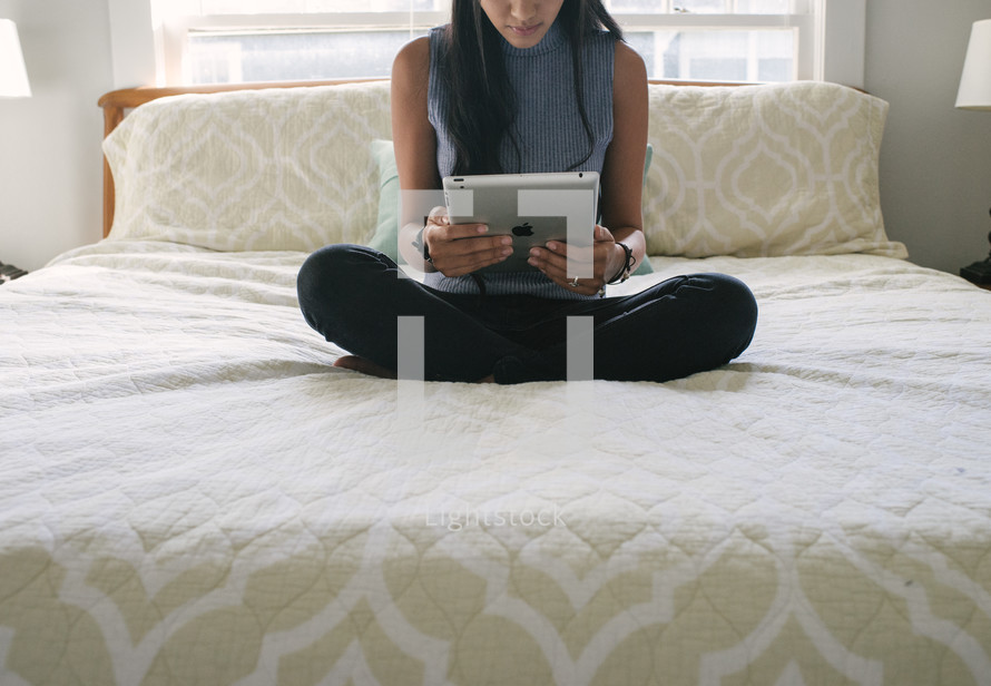 A teen girl sitting on her bed and reading an electronic tablet.