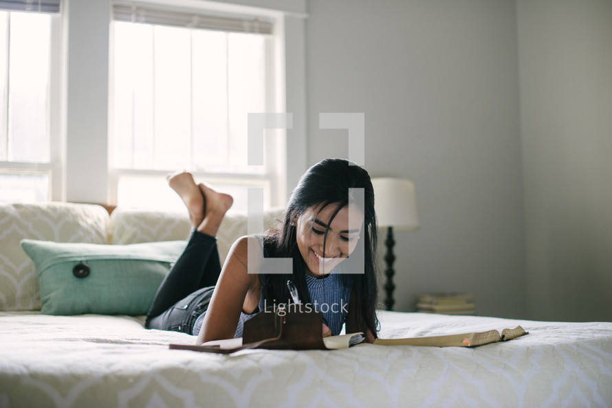 A smiling teen girl taking notes and studying the Bible while laying on her bed.