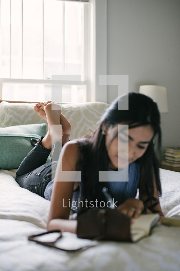 A teen girl taking notes and studying while laying on her bed.